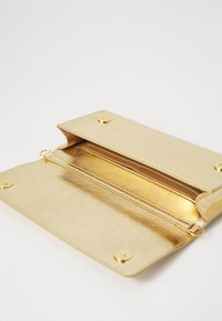 Pochette métallique dorée avec rabat ouvert, montrant le compartiment intérieur avec fermeture éclair et boutons magnétiques, sur fond blanc.