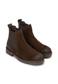 Une paire de bottes Chelsea en daim marron foncé avec des panneaux élastiques sur les côtés et des languettes de tirage sur fond blanc.