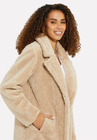 Manteau en fausse fourrure beige avec un col cranté, des poches latérales et une texture douce. Présente une coupe décontractée et des coutures détaillées.