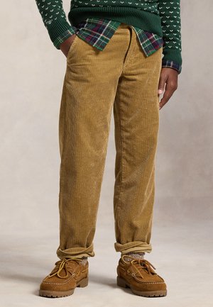 Personne portant un pantalon en velours côtelé beige retroussé aux chevilles, des chaussures marron à lacets, un pull vert et une chemise à carreaux, les mains dans les poches.