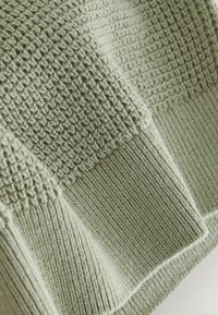 Pulover tricotat în verde murdar, cu un model texturat și tivu ribdat. Țesătura are o textură moale, ușoară, cu un ușor luciu.