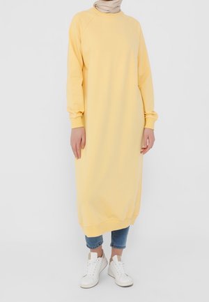 Gul sweatshirtklänning med långa ärmar, ribbade manschetter och en avslappnad passform. Buren över blå jeans och matchad med vita sneakers.