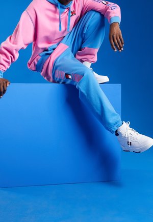 Osoba ubrana w różowo-niebieską bluzę z kapturem i pasujące spodnie dresowe, siedząca na niebieskim bloku, w białych sneakersach za kostkę na niebieskim tle.