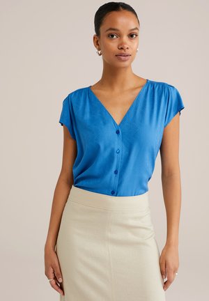 Blouse bleue à manches courtes et décolleté en V avec des boutons décoratifs et une texture subtile, associé à une jupe beige clair taille haute.