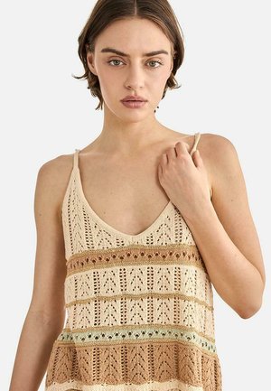 Joven con cabello corto y castaño, lleva un top sin mangas de punto con patrones horizontales en beige, marrón y verde menta, sujetándose la correa en el hombro.