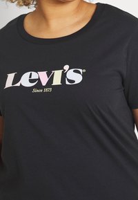 Zwarte T-shirt met pastelgekleurde "Levi's Sinds 1873" logo, gedragen door een persoon met krullend haar, weergegeven van de nek tot midden torso.