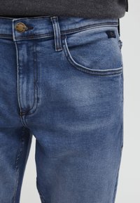 Jeans de mezclilla azul con un diseño clásico de cinco bolsillos, lavado claro, ligero desgastado y cierre de botón de metal con detalle de marca.
