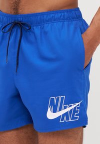 Shorts de bain bleus avec une taille élastique et un cordon de serrage, présentant un logo Nike blanc en bas à droite, un tissu lisse et des poches latérales.