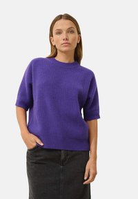 Fioletowy sweter z dzianiny z krótkimi rękawami, okrągłym dekoltem, prążkowaną fakturą, luźnym krojem, zestawiony z ciemną spódnicą.