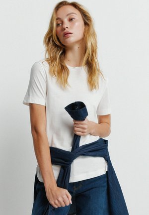 Młoda kobieta o długich blond włosach ma na sobie białą koszulkę i ciemnoniebieskie dżinsy, związaną wokół talii pasującą koszulę, trzymając za jej rękaw.