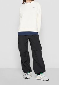 Cream sweatshirt with a small logo, layered over a dark blue top. Black cargo pants with pockets and loose fit. White and green sneakers.