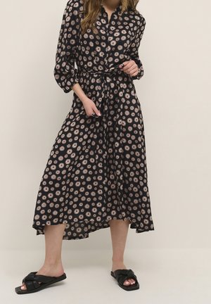 Woman wearing a black midi dress with pink floral pattern, three-quarter sleeves, tied waist, and black open-toe flat sandals.