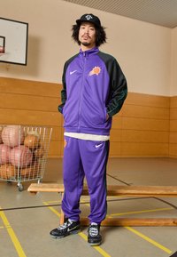 Fialová Nike tepláková súprava s čiernymi rukávmi a zelenými akcentmi, s logom basketbalu. Čierne tenisky s textúrovaným dizajnom. Drevená lavička v telocvični.