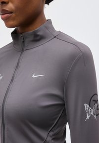 Κοντινό πλάνο ατόμου που φορά γκρι ζακέτα Nike με φερμουάρ, με γραφικό πεταλούδας και το "72" στο μανίκι.