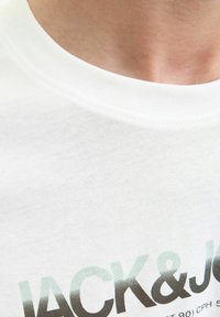 Balts T-krekls ar apaļu kakla izgriezumu un mīkstu tekstūru. Priekšpusē ir zīmola nosaukums "JACK & JONES" gradientā pelēkā un melnā krāsā.