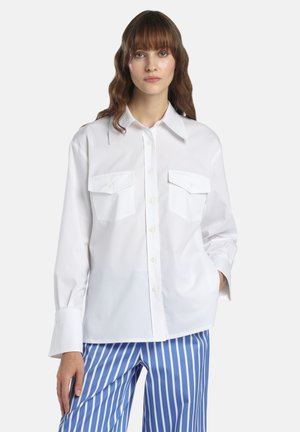 Vrouw die een oversized wit overhemd met twee borstzakken draagt en blauw-wit gestreepte broek met verticale strepen, staand met één hand in de zak.