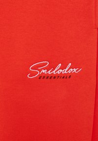 Červená bavlněná látka s vyšitým logem značky "Smilodoux ESSENTIALS" v bílé a černé barvě. Hladká textura s ležérním designem.