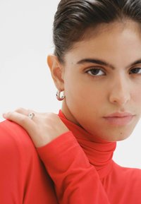 Jersey de cuello alto rojo con una textura suave, que presenta un primer plano de aretes de aro plateados y un anillo minimalista en una mano descansando sobre el hombro.
