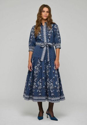 Donna che indossa un abito midi blu con fantasia floreale, maniche a tre quarti, cintura annodata in vita, calze nere e tacchi alti a punta.