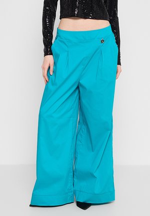 Pantaloni turchese a gamba larga con pieghe sul davanti e una spilla a forma di stella, indossati con un crop top nero a maniche lunghe con paillettes.