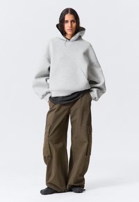Giovane con lunghi capelli indossa una felpa oversize grigia chiara, pantaloni cargo scuri e scarpe nere, in piedi con le mani in tasca.