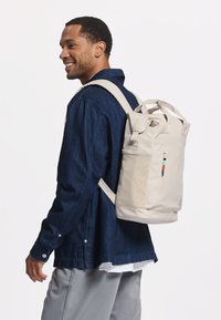 Helle beigefarbene Rucksack mit Rolltop-Design, strukturiertem Stoff und einer Seitentasche mit Reißverschluss. Verstellbare Träger und farbenfrohe Akzente sind sichtbar.