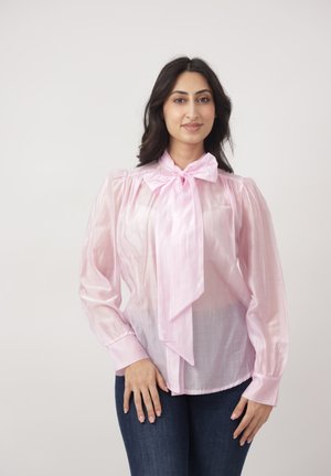 Vrouw met donker haar, gekleed in een doorschijnende roze blouse met een grote strik en een donkere spijkerbroek, staand tegen een effen lichte achtergrond.