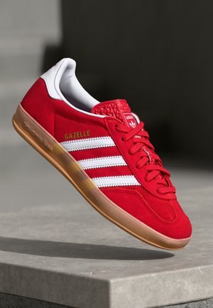 Baskets rouges Adidas Gazelle avec des bandes blanches, une semelle en gomme et un marquage doré, présentées en angle sur une surface en béton avec un fond sombre.