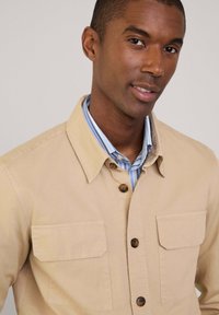 Beige Hemd mit Knopfverschluss und zwei Brusttaschen, aus Baumwollmischung, mit Kragen und durchgehender Knopfleiste. Das Innenshirt hat blaue Streifen.