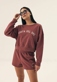 Bordowy, krótkotrwały sweatshirt z napisem "COSTA DEL SOL" w białym kolorze. W komplecie z pasującymi szortami z detalem logo. Gładki materiał, luźny krój.