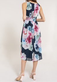 Femme de dos, portant une robe sans manches bleu marine avec un grand motif floral rose, blanc et bleu, et des sandales à talons légers rose pâle.