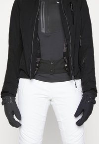 Personne portant une veste d'hiver noire, des gants noirs et un pantalon de neige blanc, visible du milieu du torse aux mains sur fond blanc.