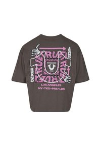 Brūns cropped t-krekls ar rozā un baltu grafisko dizainu uz muguras, kurā ir redzamas skeleta rokas, teksts un izteikts raksts.