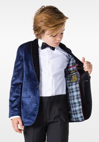 Tumes sineplict tuxedo jakk musta shawl-lapelliga, millel on ruuduline sinine sisemus ja humoorikas brodeeritud tsitaat. Valge ülikonna särk ja lips.