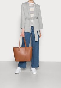 Persona vistiendo una chaqueta gris con cinturón, una camisa de rayas, jeans de pierna ancha azules, zapatillas blancas y sosteniendo un gran bolso de cuero marrón.