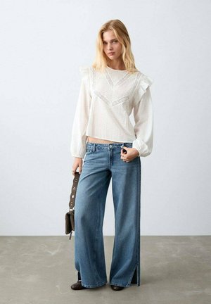 Blusa blanca con mangas con volantes y detalles de encaje, combinada con pantalones vaqueros de pierna ancha en azul con aberturas laterales. Bolso marrón. Fondo claro.