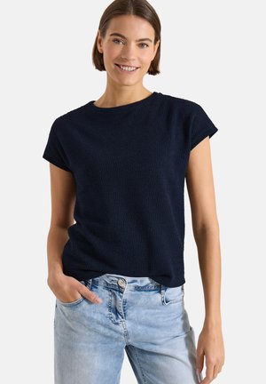Jeune femme souriante, portant un haut texturé bleu marine à manches courtes et un jean bleu clair, une main dans la poche, sur un fond uni.
