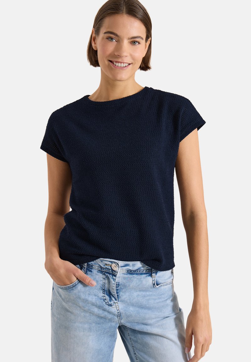 Jeune femme souriante, portant un haut texturé bleu marine à manches courtes et un jean bleu clair, une main dans la poche, sur un fond uni.