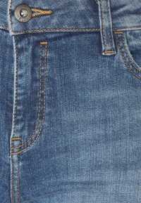 Primer plano del bolsillo de unos vaqueros de mezclilla azul con costuras naranjas, botón metálico y detalle de la presilla del cinturón.