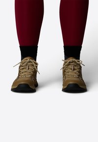 Brune tursko med teksturert overflate, med lysebrune snører og svarte såler, iført med mørke sokker og røde leggings.