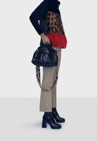 Černá lesklá kabelka s tahem na horní straně a koženým páskem se vzorem leopardí kůže. Modelka má na sobě červeno-černo-hnědou fleecovou vestu se zapínáním na zip a béžové kalhoty.