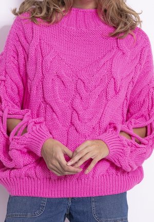 Pull tricot épais rose vif avec motifs torsadés et manches découpées nouées, porté avec un jean bleu, mains doucement jointes à la taille.