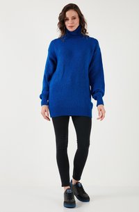 Blauwe oversized turtleneck trui in textuur gebreid met geribbelde manchetten en zoom, gecombineerd met zwarte skinny jeans en zwarte sneakers.
