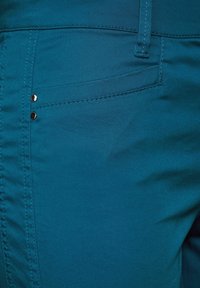 Tyrkysová látka na kalhoty s hladkou texturou. Má přední kapsu, dva dekorativní stříbrné knoflíky a kontrastní šití.