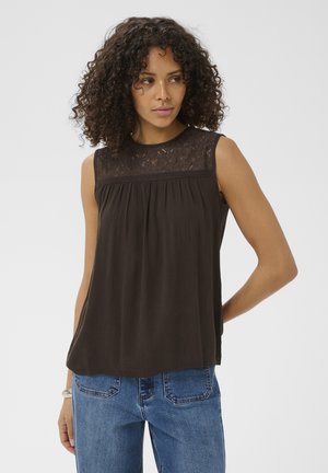 Žena s kučeravými vlasmi, ktorá má na sebe čierny čipkovaný top bez rukávov a modré džínsy, stojí s jednou rukou za chrbtom.