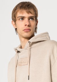 Beige hettegenser med en myk tekstur, utstyrt med snøring i hetten og et brodert logo-patch. Elegant, minimalistisk design med lette sømdekorasjoner.