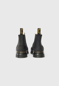 Par de botas de tornozelo de couro preto com solas grossas e costura amarela, vistas de trás sobre um fundo branco.