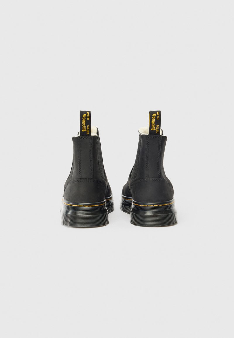 Une paire de bottines en cuir noir avec des semelles épaisses et des coutures jaunes, vues de l'arrière sur un fond blanc.