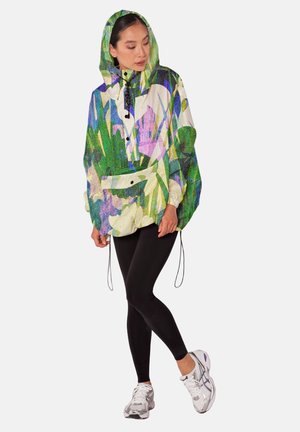 Vrouw draagt een kleurrijk, met bloemen bedrukt hoodiejack, zwarte leggings en witte hardloopschoenen, staand met gekruiste benen.