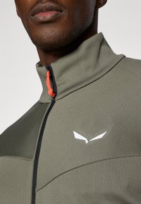 Herren-Jacke mit Reißverschluss aus gedämpftem olivgrünem Stoff. Verfügt über einen hohen Kragen, einen schwarzen Reißverschluss und ein weißes Logo auf der Brust. Heller orangefarbener Zipper-Akzent.